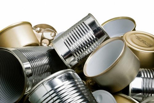 A close-up shot of multiple aluminium and tin cans, some with pull-tab lids and others with sealed tops, piled together on a surface, likely in an outdoor or industrial setting. The cans vary in colour, with some painted in metallic gold and others in silver, exhibiting different finishes such as glossy or matte. The image shows the cans stacked and leaning at various angles, with some partially obscured. The background is neutral and out of focus, emphasizing the cans' reflective surfaces and textures. This arrangement suggests waste or recycling materials, consistent with rubbish removal services provided by Office Clearance Ickenham. The scene captures the typical appearance of discarded food or beverage containers, which are often collected during rubbish collection or clearance operations in the Ickenham area, a detail subtly supporting local SEO signals for waste disposal services.